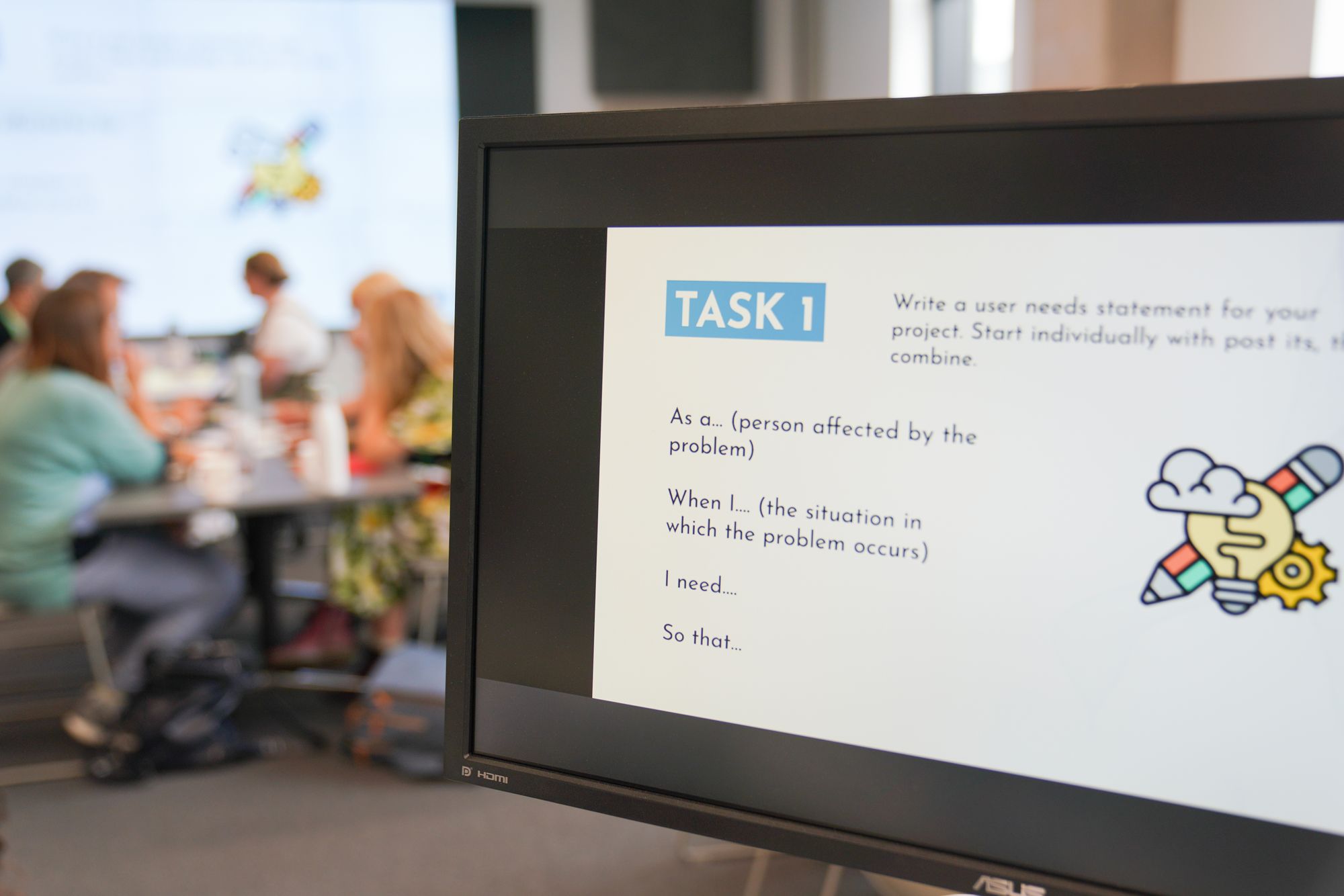 A presentation on a computer screen that says ‘Task 1’. Blurred groups of people collaboratively working at desks in the background.