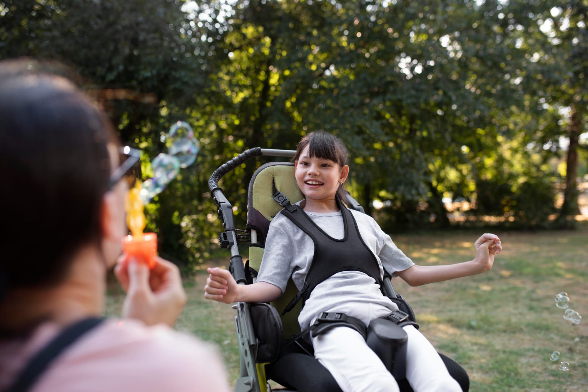 A mother blows bubbles for her smiling young daughter, who is sitting in a wheelchair.