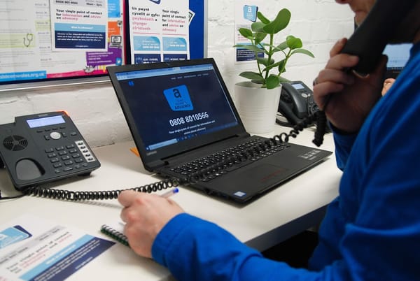 Advisor for Gwent Access to Advocacy helpline talking on the phone while taking notes with a pen and paper at a desk. 